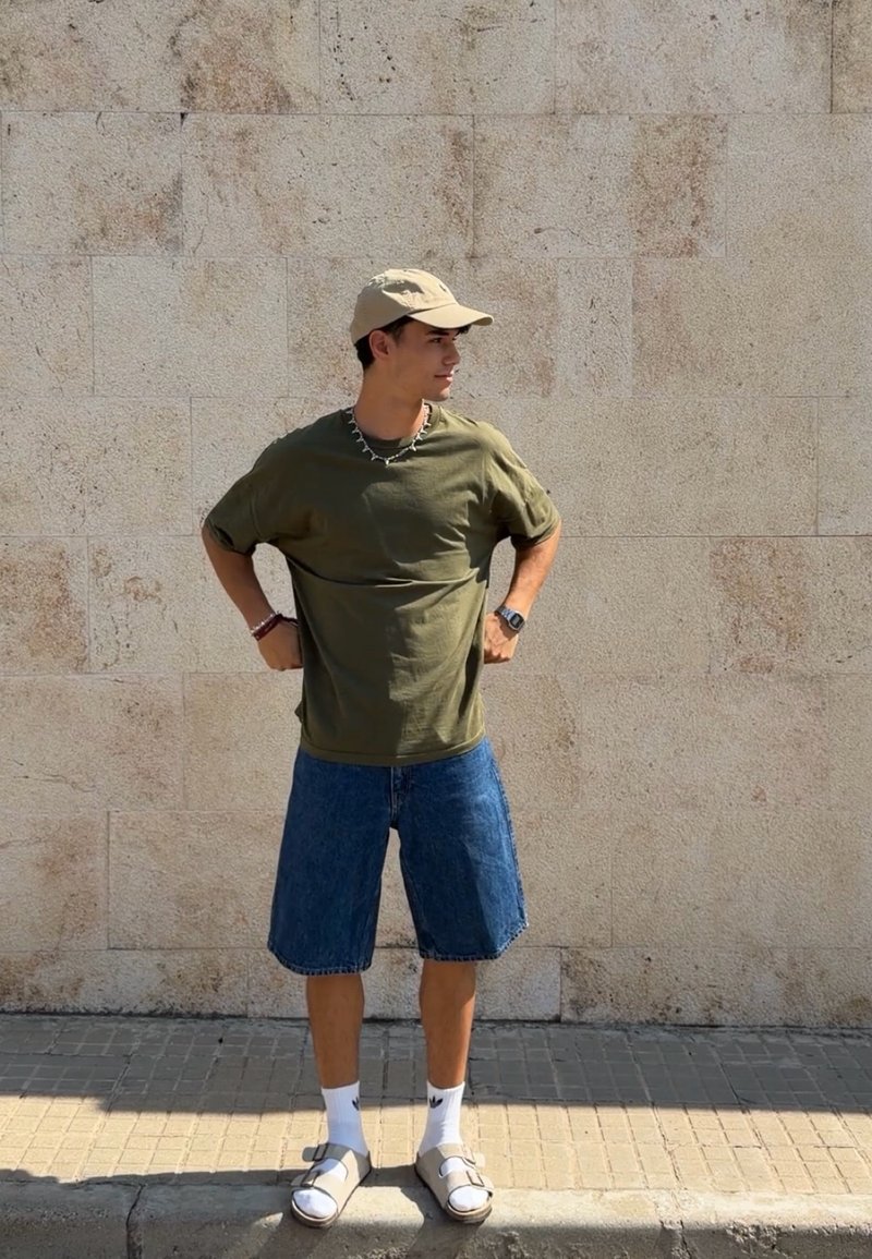 Jeune homme portant un t-shirt vert olive, un short en jean, une casquette beige, des chaussettes blanches et des sandales, debout contre un mur en pierre beige.