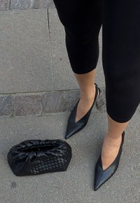 Czarne skórzane buty na wysokim obcasie typu slingback z ostrym noskiem i w kształcie litery V wycięciami, w połączeniu z plecioną czarną torebką kopertową, na szarej kostce brukowej.