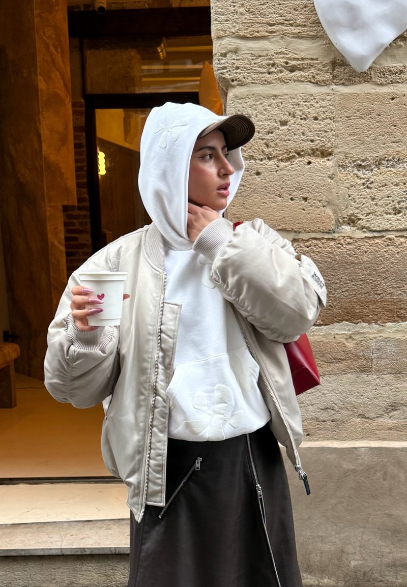 Frau in beigefarbener Bomberjacke und weißem Kapuzenpullover, die eine Kaffeetasse hält, seitlich blickend, steht an einer Steinmauer neben einer offenen Tür.