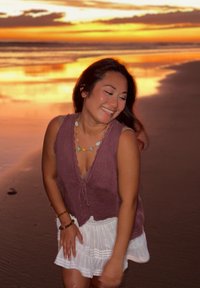 Femme souriante aux cheveux foncés, portant un haut violet sans manches et une jupe blanche, debout sur une plage au coucher du soleil.