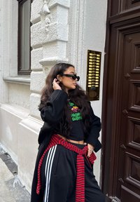 Svart oversized kappa, svart croppad t-shirt med grön tryck, svarta sweatpants med vita ränder, röd randig midjerem, mörka solglasögon.