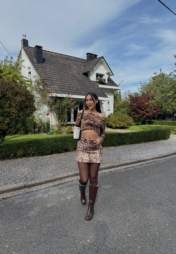 Joven mujer con falda marrón estampada y top con botas camina por una calle sosteniendo una taza, con una casa blanca y un jardín al fondo.