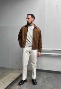 Καφέ σουέτ ζακέτα με φερμουάρ, φορεμένη πάνω από ένα ανοιχτό γκρίζο φούτερ, συνδυασμένη με λευκά παντελόνια ίσια�ς γραμμής και μαύρα δερμάτινα παπούτσια.