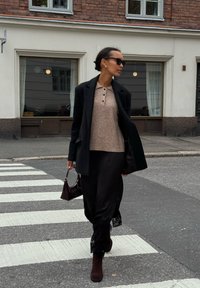 Cappotto nero, maglione corto a maniche corte color marrone chiaro con bottoni, gonna nera con dettagli in pizzo, stivali con tacco marroni e una piccola borsa scura.