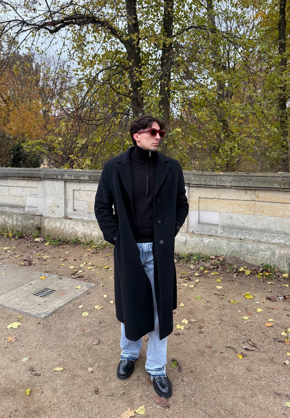 Μαύρο μακρύ παλτό πάνω από μαύρο ζιβάγκο, συνδυασμένο με ανοιχτ�ό γαλάζιο τζιν. Μαύρα παπούτσια και ροζ γυαλιά ηλίου. Περιτριγυρισμένος από φθινοπωρινά φύλλα.