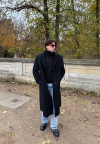 Μαύρο μακρύ παλτό πάνω από μαύρο ζιβάγκο, συνδυασμένο με ανοιχτό γαλάζιο τζιν. Μαύρα παπούτσια και ροζ γυαλιά ηλίου. Περιτριγυρισμένος από φθινοπωρινά φύλλα.