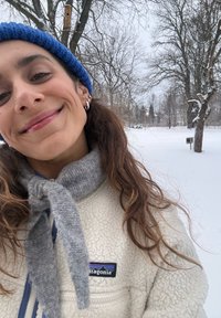 Veste en polaire blanche avec une fermeture éclair et un logo, écharpe en maille grise, et bonnet bleu. Arrière-plan enneigé avec des arbres nus.