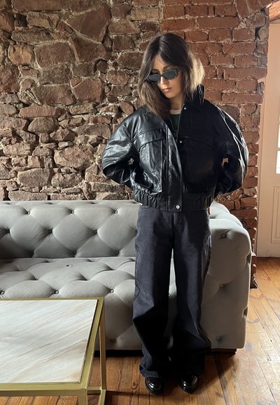 Chaqueta bomber corta de cuero negro con grandes bolsillos, combinada con jeans de mezclilla oscuros de cintura alta, contra un fondo de pared de ladrillo.