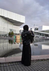 Person som bär en svart läderjacka med fransar, vida svarta byxor och huva, som tar ett foto nära en reflekterande vattenspegel och moderna byggnader.