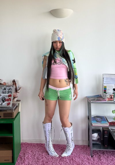 Joven mujer con cabello largo de pie llevando un crop top rosa, pantalones cortos verdes, botas blancas hasta la rodilla con cordones, sombrero estampado y chaqueta sobre un hombro.