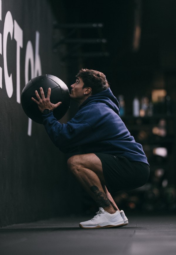 Mann in blauem Kapuzenpullover und schwarzen Shorts hockt und hält einen Medizinball, bereit, ihn gegen eine Gymnastikwand zu werfen.