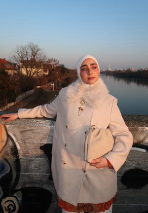 Joven mujer con abrigo blanco y hijab apoyada en la barandilla de un puente cubierto de grafitis, sosteniendo un bolso beige, con un río y casas al fondo al atardecer.