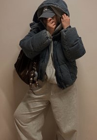 Geacă puf din denim închis la culoare cu glugă, hanorac gri dedesubt, pantaloni de trening gri, geantă maro de umăr cu accente de breloc, și șapcă gri.