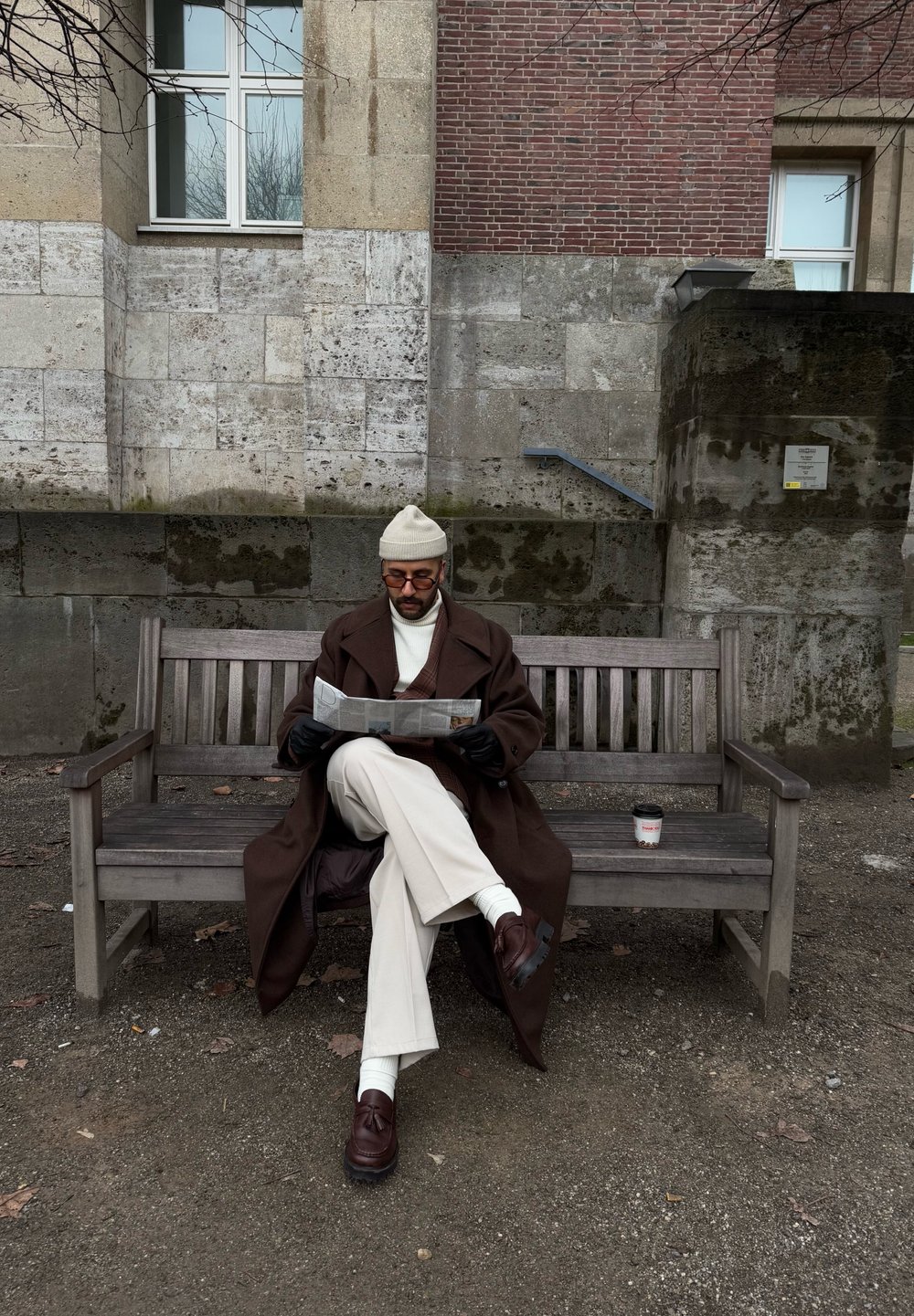 Καφέ παλτό, κρεμ ζιβάγκο, μπεζ σκουφί, μαύρα γάντια, λευκά παντελόνια και καφέ παπούτσια. Κρατάει μια εφημερίδα σε ένα ξύλινο παγκάκι.