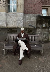 Καφέ παλτό, κρεμ ζιβάγκο, μπεζ σκουφί, μαύρα γάντια, λευκά παντελόνια και καφέ παπούτσια. Κρατάει μια εφημερίδα σε ένα ξύλινο παγκάκι.