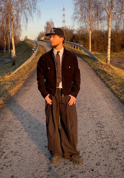 Joven con chaqueta negra, pantalones anchos y gorra, de pie en una carretera iluminada por el sol cerca de árboles de abedul al atardecer, mirando hacia un lado.