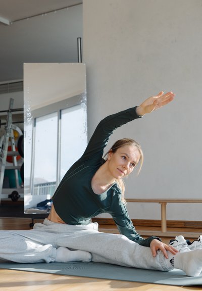 Figura femenina con un top de manga larga verde oscuro y pantalones de chándal grises realizando un estiramiento lateral sobre una esterilla de yoga en un gimnasio con espejos.