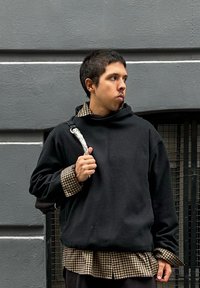 Czarny, oversize hoodie z wysokim kołnierzem, nałożony na brązową koszulę w kratę. Trzyma pasek z srebrnym akcentem. Szare tło.