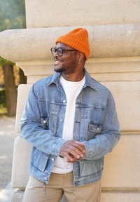 Lys blå denimjakke med to brystlommer, båret over en hvid t-shirt. Orange ribbet hue og beige bukser. Struktureret stenvæg i baggrunden.