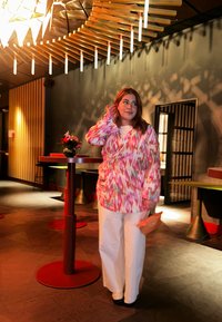 Färgglad, mönstrad wrap-overdel med rosa, orange och gröna nyanser; vita vida byxor; liten beige väska; svagt upplyst modern inredning.