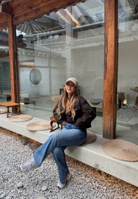Jeune femme en vêtements décontractés et casquette assise sur un banc en plein air près d'un mur en verre, sur un sol en gravier, avec des piliers en bois rustique.
