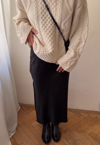 Asmuo, apsirengęs kreminės spalvos megztiniu su iškeltu raštu, juoda midi ilgio sijonu, juodomis botais ir juoda per petį nešama rankine, stovi ant medinių grindų šalia baltos užuolaidos.