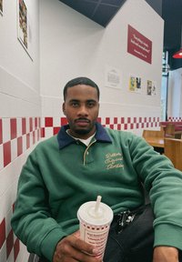 Grön och mörkblå långärmad pikétröja med gula detaljer. Håller en randig mugg med sugrör. Röd- och vitkaklad vägg i bakgrunden.