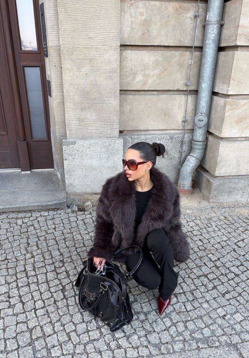 Femme portant un manteau sombre en fourrure et des lunettes de soleil, agenouillée sur un trottoir pavé, tenant un sac à main noir, près d'un bâtiment en pierre et d'une porte.
