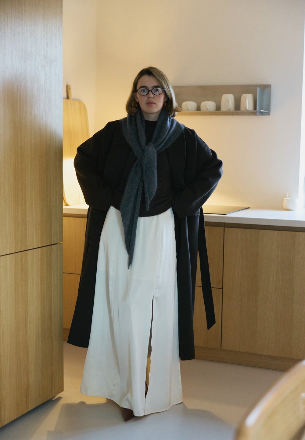 Prevelik črni plašč, sivi šal, temen zgornji del in bele hlače s stranskimi razporkami, ki poudarjajo sproščen kroj, mehke teksture in nevtralne barve.
