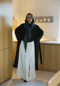 Prevelik črni plašč, sivi šal, temen zgornji del in bele hlače s stranskimi razporkami, ki poudarjajo sproščen kroj, mehke teksture in nevtralne barve.