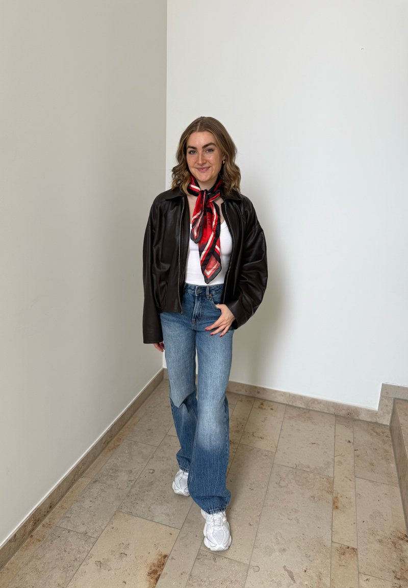 Giovane donna in piedi in casa che indossa una giacca nera, una maglietta bianca, una sciarpa rossa, jeans blu e scarpe da ginnastica bianche, sorridente verso la fotocamera.