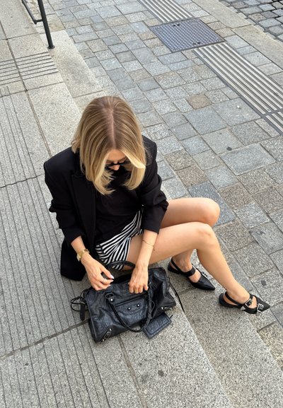 Bolso de mano de cuero negro con superficie texturizada, herrajes plateados y asas dobles; combinado con una falda a rayas y zapatos negros. Pasos de concreto y pavimento de fondo.