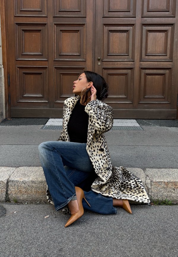 Mujer sentada en el bordillo de la calle, con un abrigo largo de estampado, vaqueros azules, tacones altos marrón claro, y ajustándose el pendiente cerca de una puerta grande de madera.