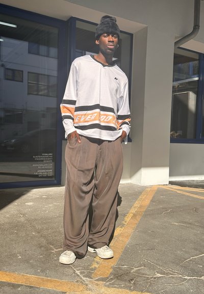 Camisa de malla blanca de manga larga con rayas naranjas y negras, acompañada de pantalones grises holgados y zapatillas blancas. Gorro negro en la cabeza.