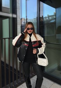 Femme portant des lunettes de soleil noires, un col roulé noir, un pantalon en cuir noir, une veste en cuir bicolore, tenant un sac à main blanc, se tenant près d'une rambarde et d'une paroi vitrée.