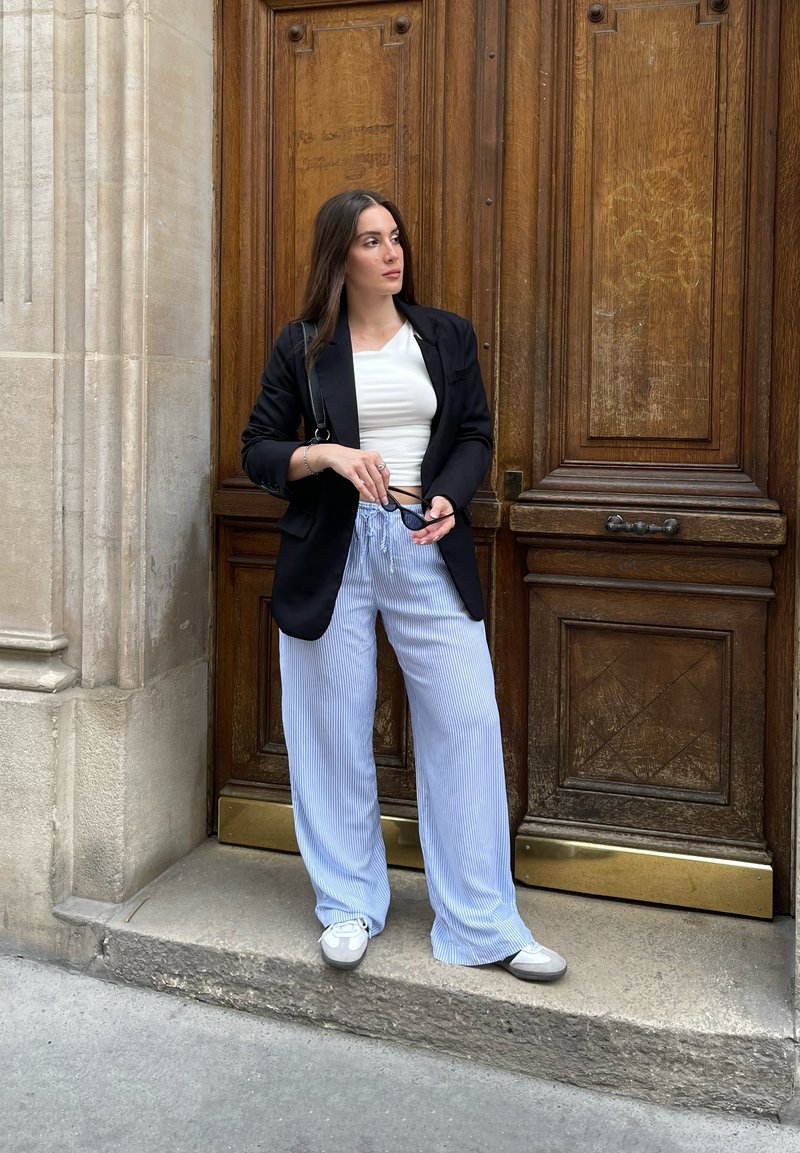 Κάτω από μια μαύρη σαλοπέτα, σε λευκό cropped μπλουζάκι, συνδυασμένα με μπλε και λευκές ριγωτές παντελόνες ευρείας γραμμής. Στέκεται σε μια πέτρινη σκάλα κοντά σε μια ξύλινη πόρτα.