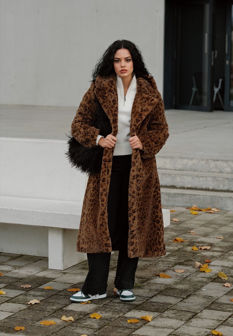 Leopard print faux fur coat with a wide collar, white zip-up sweater, black trousers, and shoes with green accents; set in an outdoor setting.