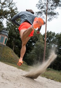Sportowiec w szarej koszulce z długim rękawem i jasnopomarańczowych szortach skacze z piaszczystej powierzchni, unosząc piasek, w pomarańczowych butach biegowych.