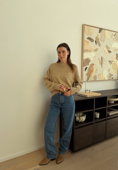 Mujer con suéter beige y jeans azules sosteniendo una pequeña cartera marrón, de pie junto a un mueble de madera oscura con arte abstracto en la pared encima.