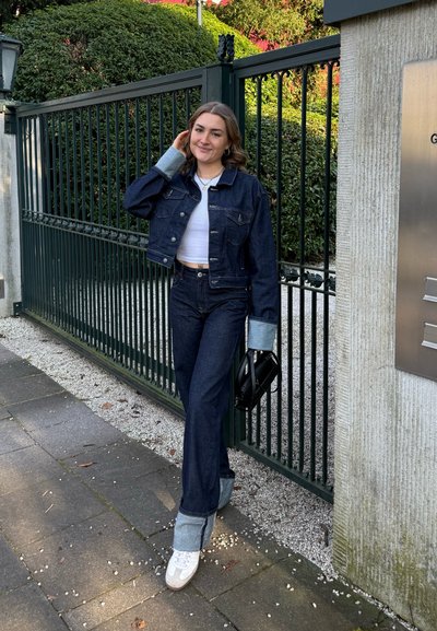 Chaqueta de mezclilla oscura con puños ampliados, combinada con unos pantalones oscuros de cintura alta. Debajo, una camiseta corta blanca y un bolso negro.