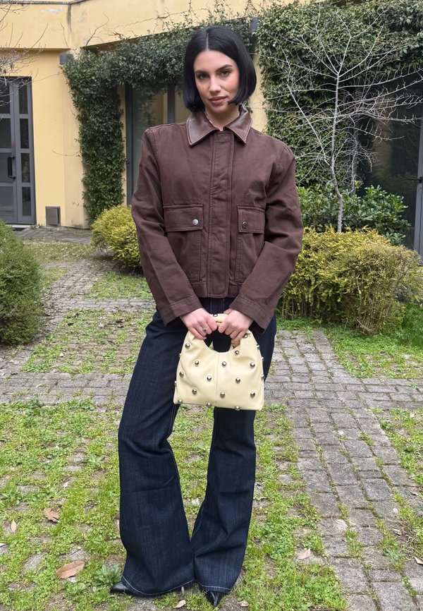 Donna con capelli corti neri che indossa una giacca marrone e jeans scuri svasati, con una borsa crema con borchie metalliche, in piedi su un sentiero di pietra.