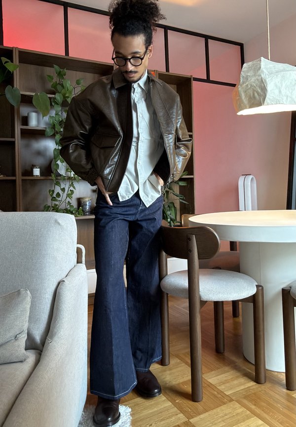 Mann mit runden Brillen, brauner Lederjacke, weißem Hemd und weit geschnittenen Jeans, der in einem Wohnzimmer neben einem Esstisch und Pflanzen steht.