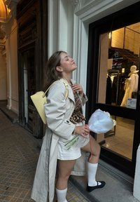 Mlada ženska v bež plašču, zgornjem delu z živalskim vzorcem, belem krilu in nogavicah drži cvetje, stoji pred butikom z zaprtimi očmi in se smeška.