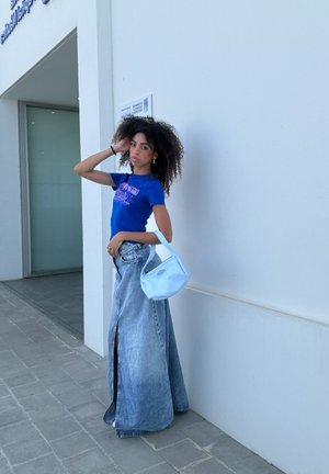 Falda de mezclilla oversized azul claro con una abertura, combinada con una camiseta gráfica azul ajustada y un pequeño bolso blanco. Texturas suaves.