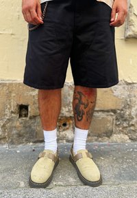Μαύρα σορτς με χαλαρή εφαρμογή, σε συνδυασμό με ανοιχτόχρωμες κάλτσες και μπεζ παπούτσια. Σημαντικός σχεδιασμός τατουάζ στο κάτω μέρος του ποδιού. Λεπτομέρεια αλυσίδας στα σορτς.