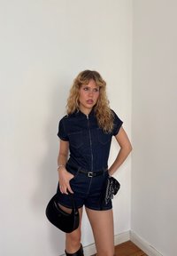 Denim haalarissa on lyhyet hihat ja edessä vetoketju, ja siinä on kaksi rintataskua sekä musta vyö. Musta käsilaukku ja kuvioitu huivi viimeistelevät ilmeen.