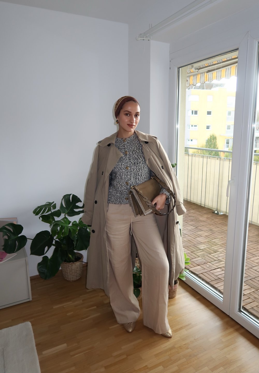 Manteau trench beige, pull en maille gris, pantalon large crème, sac à main en or texturé, chaussures beiges à bouts pointus, intérieur avec des plantes.