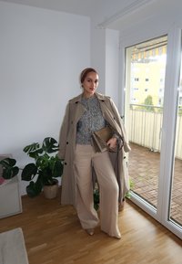 Manteau trench beige, pull en maille gris, pantalon large crème, sac à main en or texturé, chaussures beiges à bouts pointus, intérieur avec des plantes.