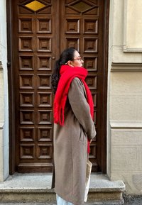 Καφέ παλτό, κόκκινο μαντήλι με φούντες και ανοιχτό μπλε παντελόνι φορεμένα από ένα άτομο που στέκεται δίπλα σε μια ξύλινη πόρτα με διαμαντόσχημα παράθυρα.