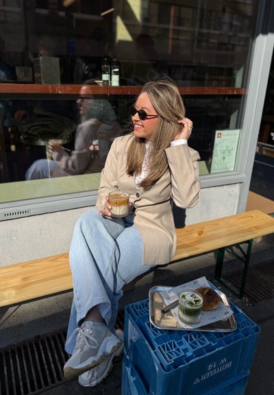 Una persona sentada sostiene una bebida helada con capas en un vaso transparente. Lleva puesto un blazer beige, pantalones azul claro y zapatillas deportivas blancas.